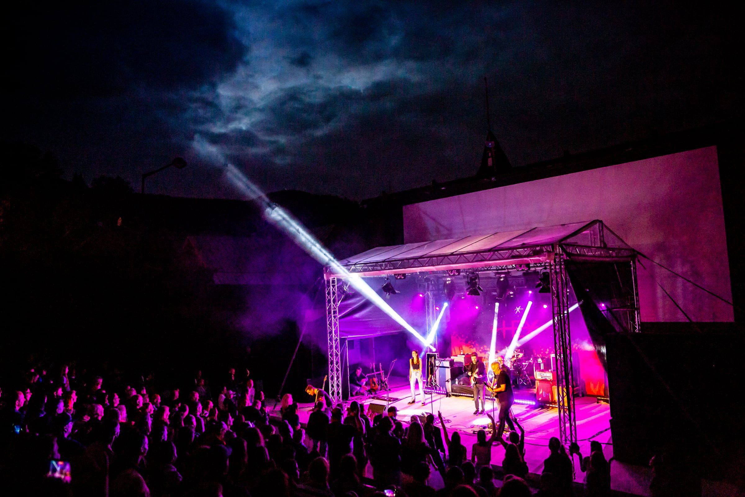 Koncert v amfiteátri v Banskej Štiavnici, pódium vo fialovom svetle a lúče reflektorov nad publikom